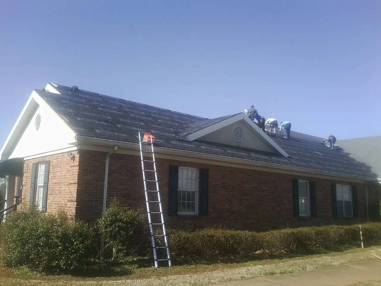 Crew tearing shingles off a brick ranch roof with a ladder set — Metal Roof Repair work by our crew in Marcy