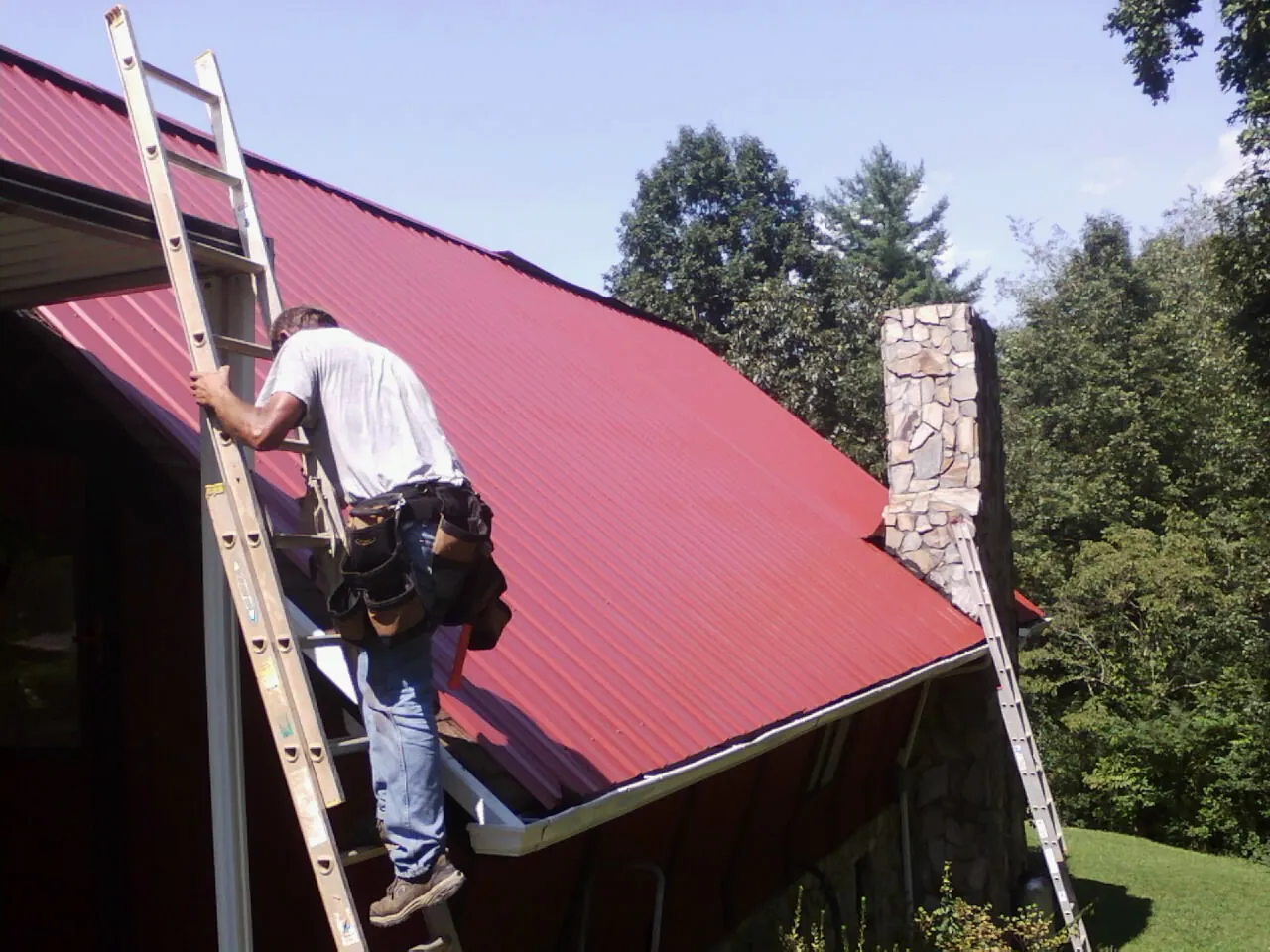 Roofer on a ladder beside a red metal roof and stone chimney — Soffit & Fascia Repair by Roofing Brothers in Marcy